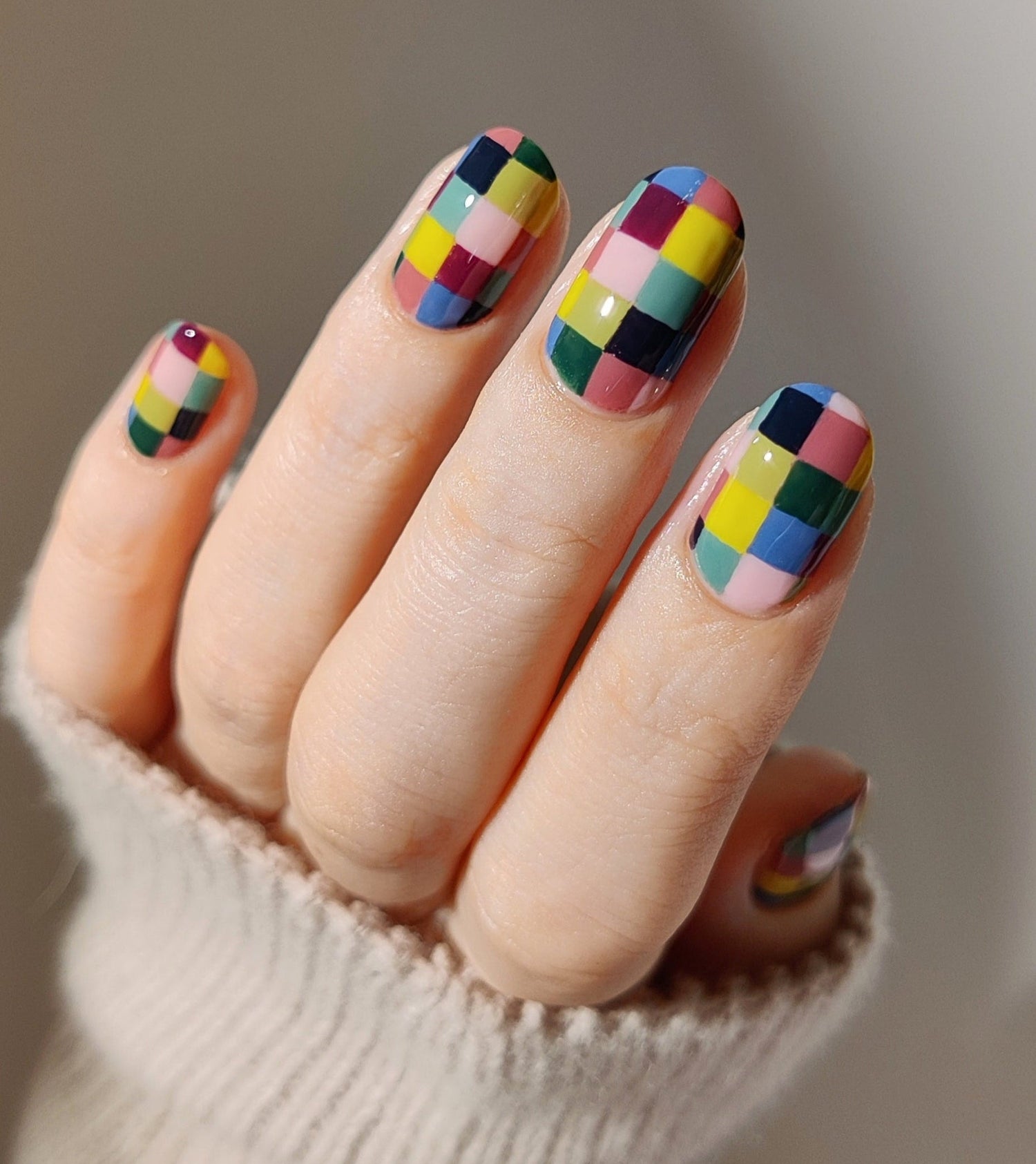Nails with colorful checkered design held by a person wearing a beige sweater.