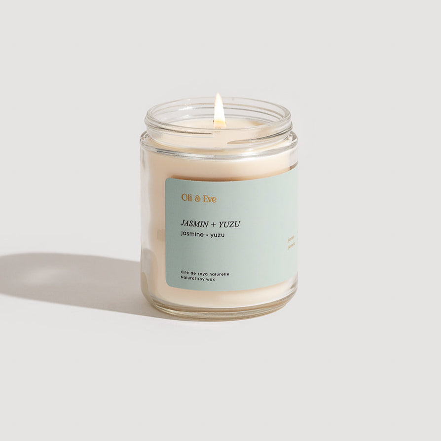 A white candle with a cotton wick, in a clear glass jar, labeled &