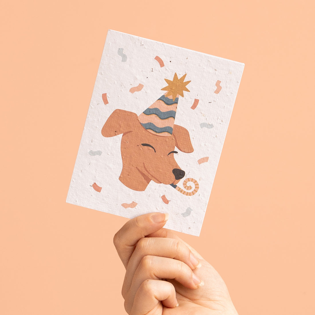 Hand holding a card with a dog wearing a party hat on a peach background