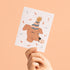Hand holding a card with a dog wearing a party hat on a peach background