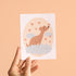 Hand holding a card with a dog illustration and heart shapes on a beige background