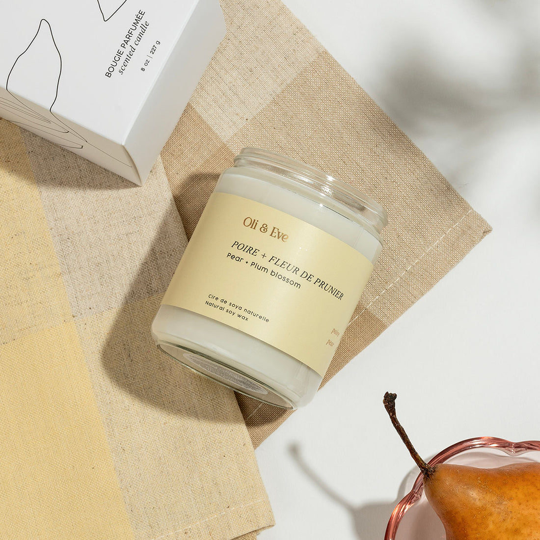 Candle jar with a label on a textured surface next to a pear